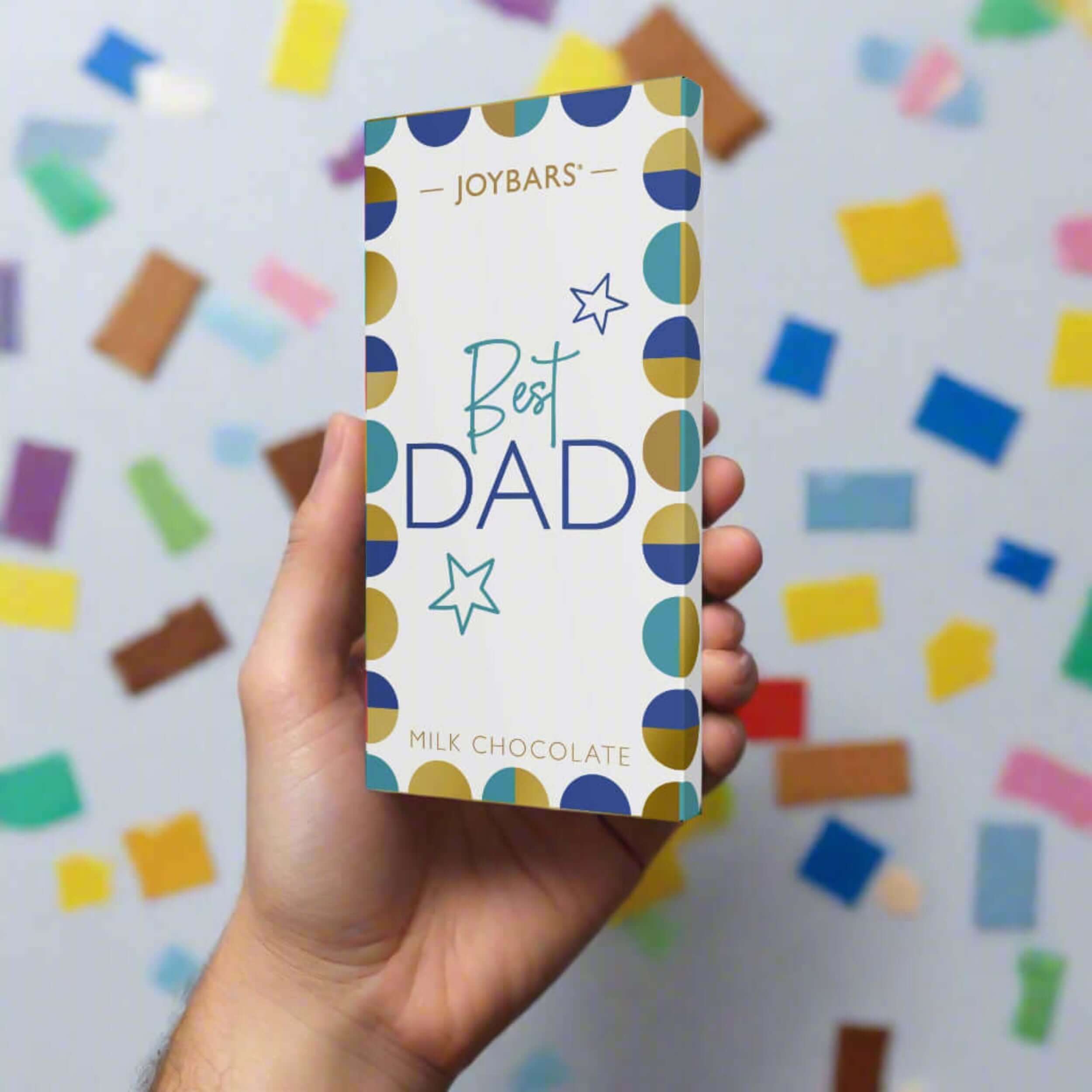 Hand holding a chocolate bar package labeled 'Best Dad' against a colorful background.
