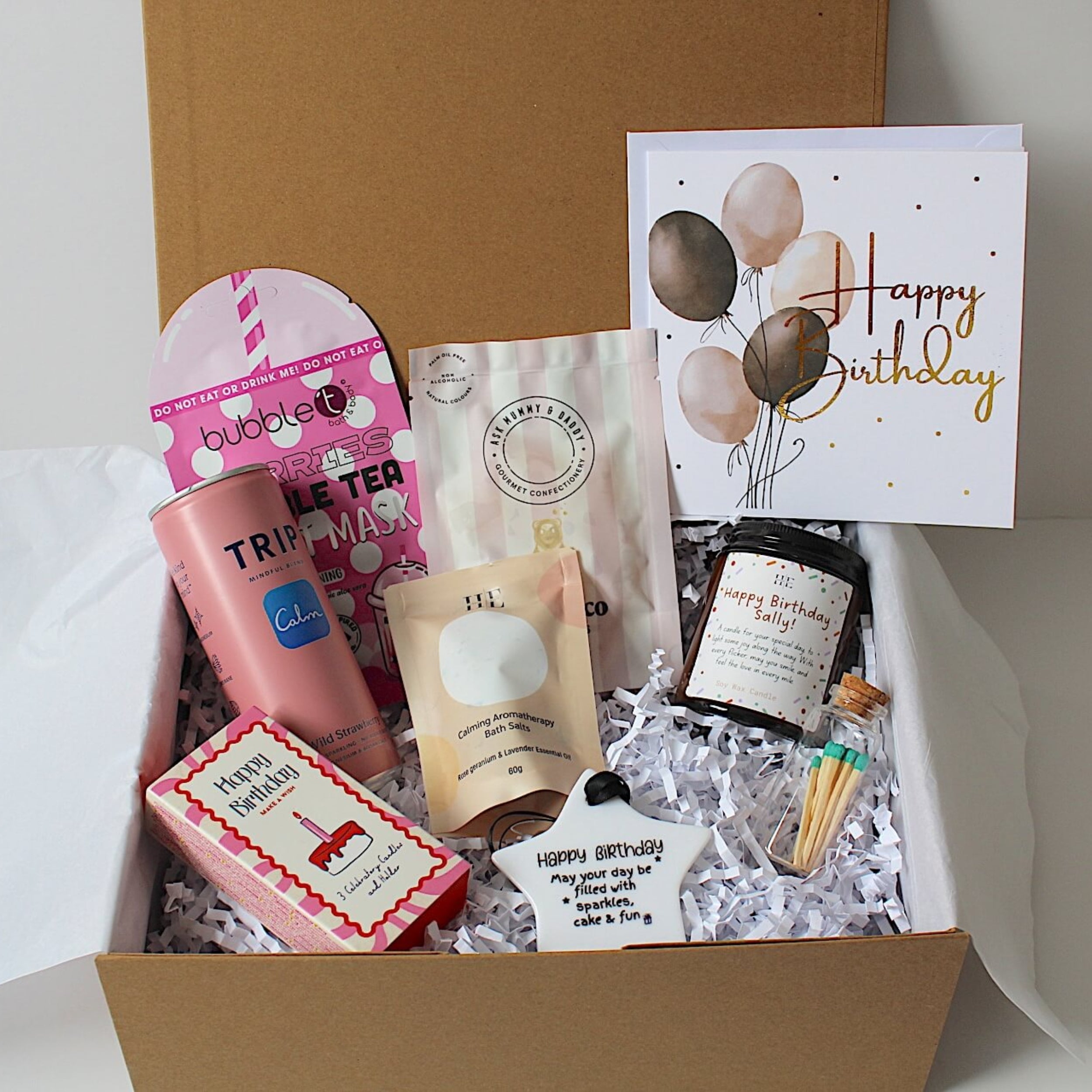 A curated birthday gift box filled with assorted treats and stationery. The box contains a pink bubble bath product, a pink tea bag pack, a beige bath & body item, a small jar, a pink and white soap bar, a white 'Happy Birthday' balloon card, and a small white 'Happy Birthday' message card, all arranged in white crinkle paper inside a brown cardboard box.