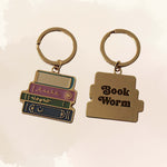 Two gold-toned book-shaped keychains on a light beige background. One keychain shows a stack of colorful books with decorative lettering, and the other is a single book with the words 'Book Worm' in black script.