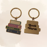 Two gold-toned book-shaped keychains on a light beige background. One keychain shows a stack of colorful books with decorative lettering, and the other is a single book with the words 'Book Worm' in black script.