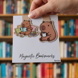 A hand holds a white card displaying two cute cartoon capybara bookmarks on a blurred library bookshelf background. The bookmarks are die-cut with brown capybara illustrations, one reading a book and one holding a mug, with the text 'Magnetic Bookmarks' and 'Boxist' printed on the card.