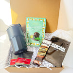 A curated gift box set in a brown cardboard box filled with white paper shred, featuring a dark gray insulated tumbler, a mint green package of KitKat Peppermint Crunch, a bag of Galaxy Natural Cacao chocolate, and a bag of Lap-Cool Charcoal Filter Coffee.