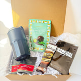 A curated gift box set in a brown cardboard box filled with white paper shred, featuring a dark gray insulated tumbler, a mint green package of KitKat Peppermint Crunch, a bag of Galaxy Natural Cacao chocolate, and a bag of Lap-Cool Charcoal Filter Coffee.