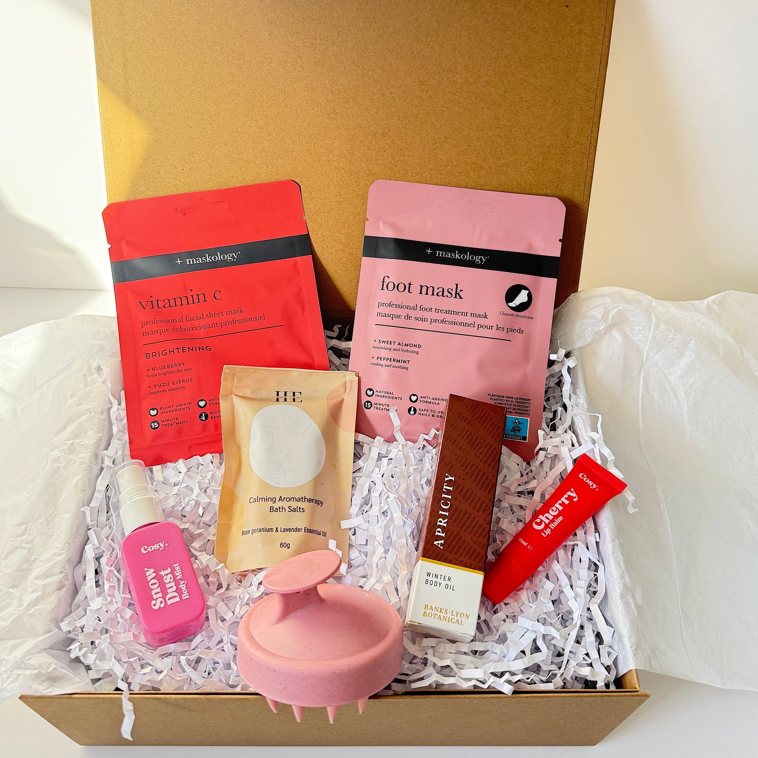 An open cardboard gift box filled with skincare products on white tissue paper. Visible items include a red and pink sheet mask pouch, a pink foot mask packet, a small pink bottle, a beige tube, a brown rectangular item, a red tube, and a pink round brush-like accessory, all arranged in white shredded paper.