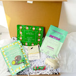 An open magnetic closure gift box filled with assorted packaged snacks and wellness items on white tissue paper. Visible items include a green Christmas elf eyes packet, a mint green detox leafy greens packet, a mint green peppermint crunch packet, a small cream-colored item, and a small red-and-green candy packet, arranged in white shredded paper.