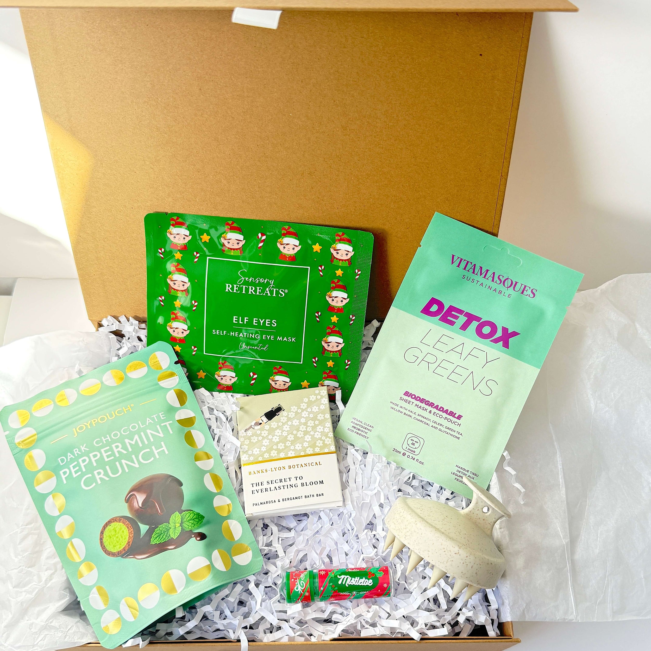 An open magnetic closure gift box filled with assorted packaged snacks and wellness items on white tissue paper. Visible items include a green Christmas elf eyes packet, a mint green detox leafy greens packet, a mint green peppermint crunch packet, a small cream-colored item, and a small red-and-green candy packet, arranged in white shredded paper.