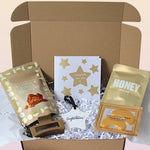 An open kraft  gift box filled with white crinkle paper, containing  Cookie & Fudge Clusters, a white card with gold stars and the words 'Congratulations you did it', a honey sheet mask, under eye patches, square claw clip and a ceramic keepsake "congratulations". 