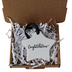 A white star-shaped ceramic ornament with 'Congratulations' written in black script, presented inside an open brown gift box filled with white crinkle paper.