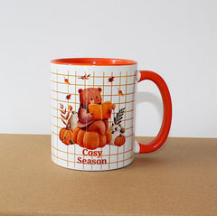 Cosy Season Mug with orange handle and inside mug, on brown sideboard with white background. The mug image is a bear sitting on a pumpkin reading. 