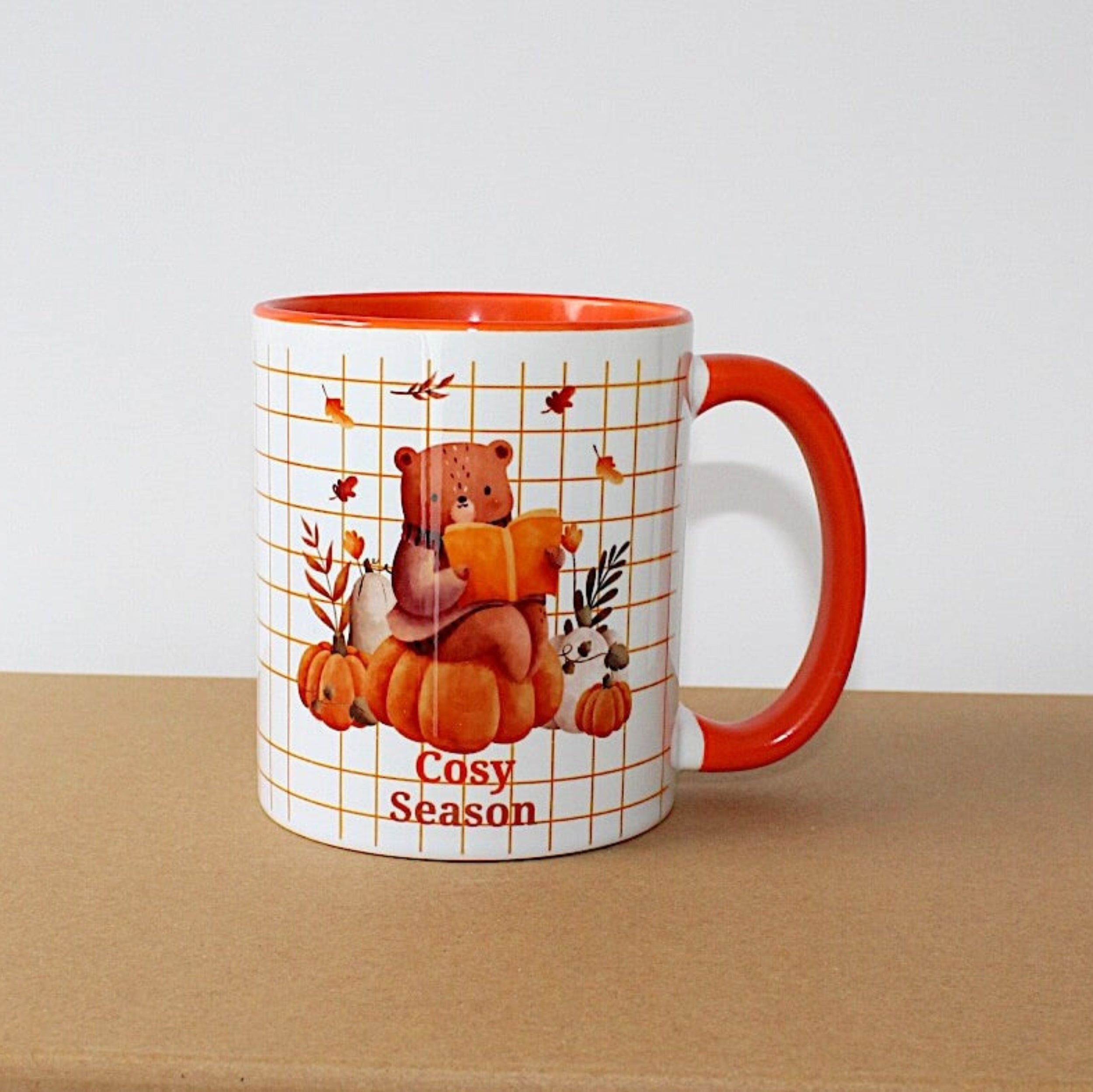 Cosy Season Mug with orange handle and inside mug, on brown sideboard with white background. The mug image is a bear sitting on a pumpkin reading. 