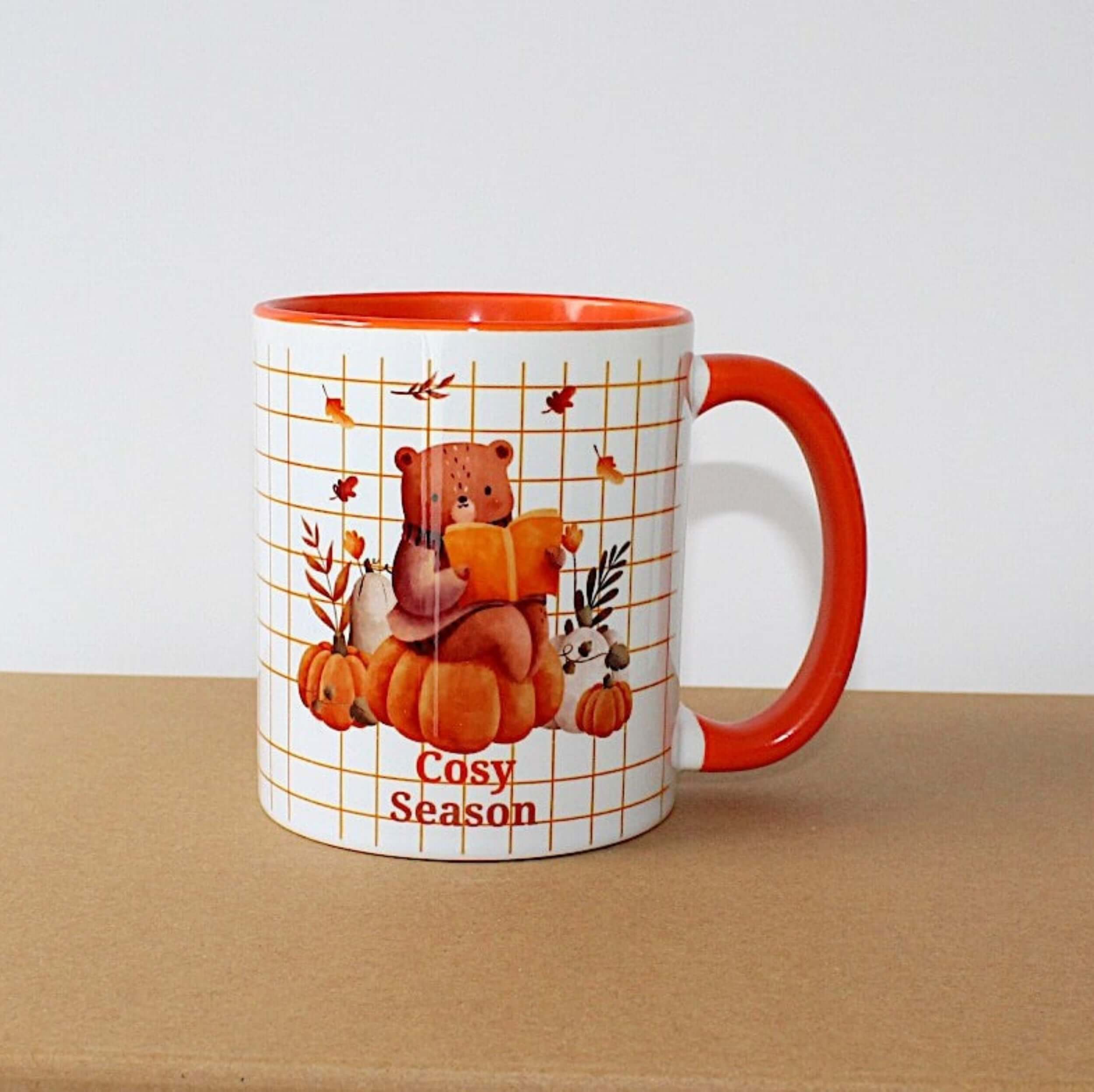 Cosy Season Mug with orange handle and inside mug, on brown sideboard with white background. The mug image is a bear sitting on a pumpkin reading. 