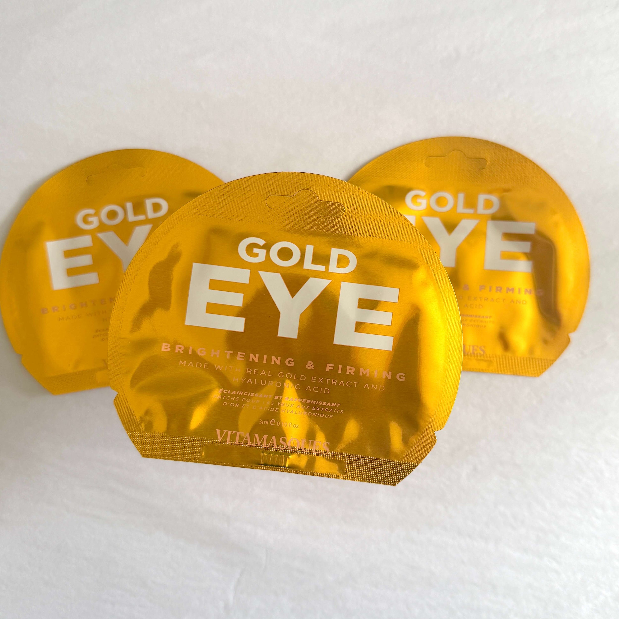 Three gold round eye mask packets with 'GOLD EYE' printed in white, arranged on a white textured fabric background.
