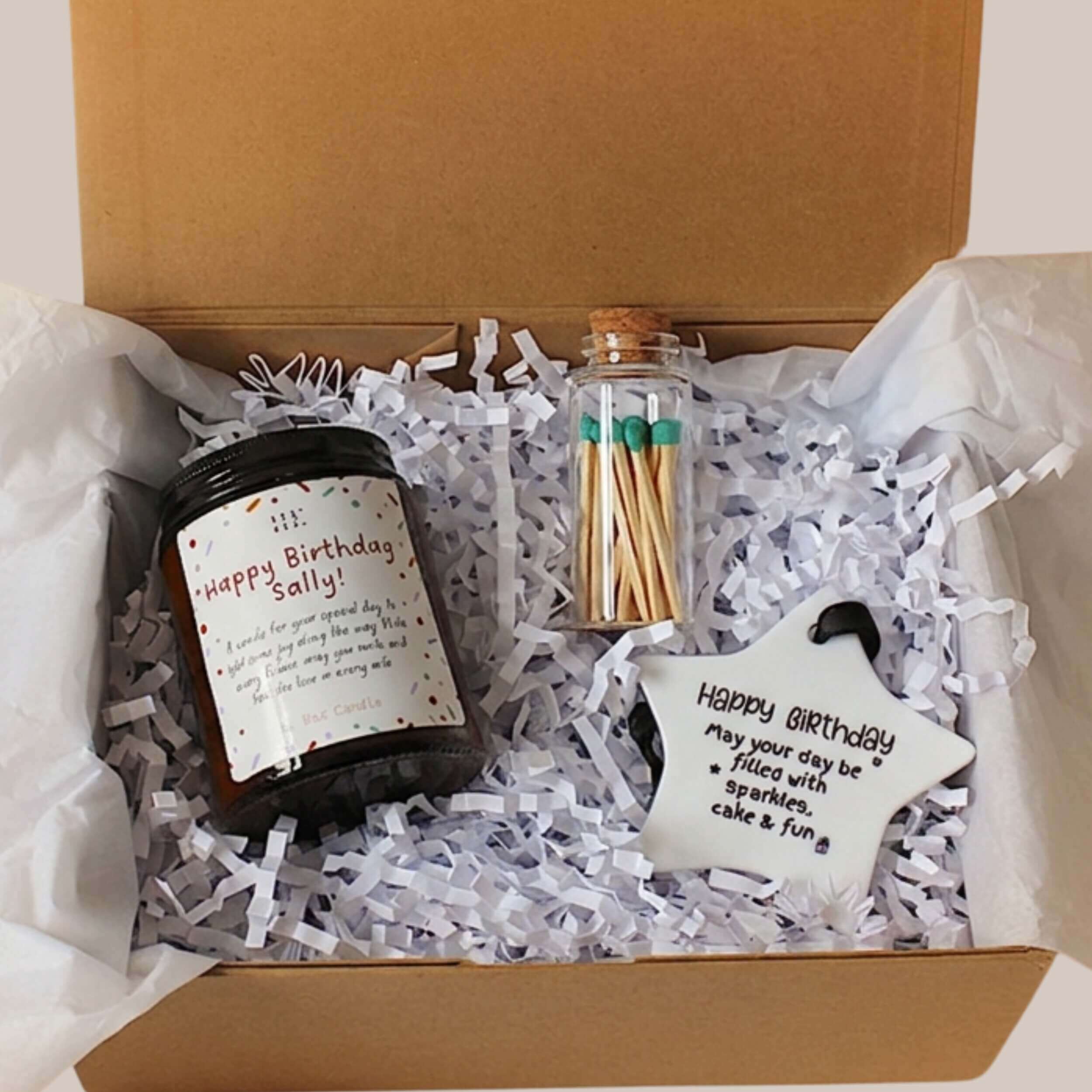 A curated birthday gift box containing a dark glass candle jar with a 'Happy Birthday Sally'  personalised label, a small glass vial of wooden matches with green tips, and a white star-shaped tag with a black script message 'Happy birthday", all arranged in white crinkle paper inside a magnetic closure gift box 