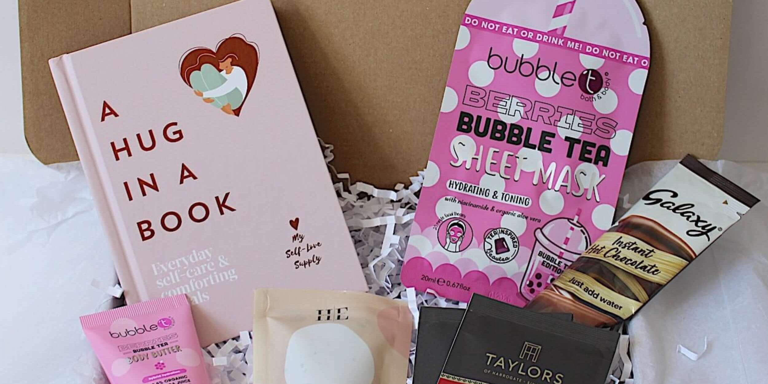 A curated gift box filled with various items including a pink hardcover book titled 'A HUG IN A BOOK', a pink bubble tea sheet mask, berries body butter, aromatherapy bath salts, two individually wrapped tea bags, and a Galaxy chocolate bar, all arranged on white tissue paper inside a gift box.