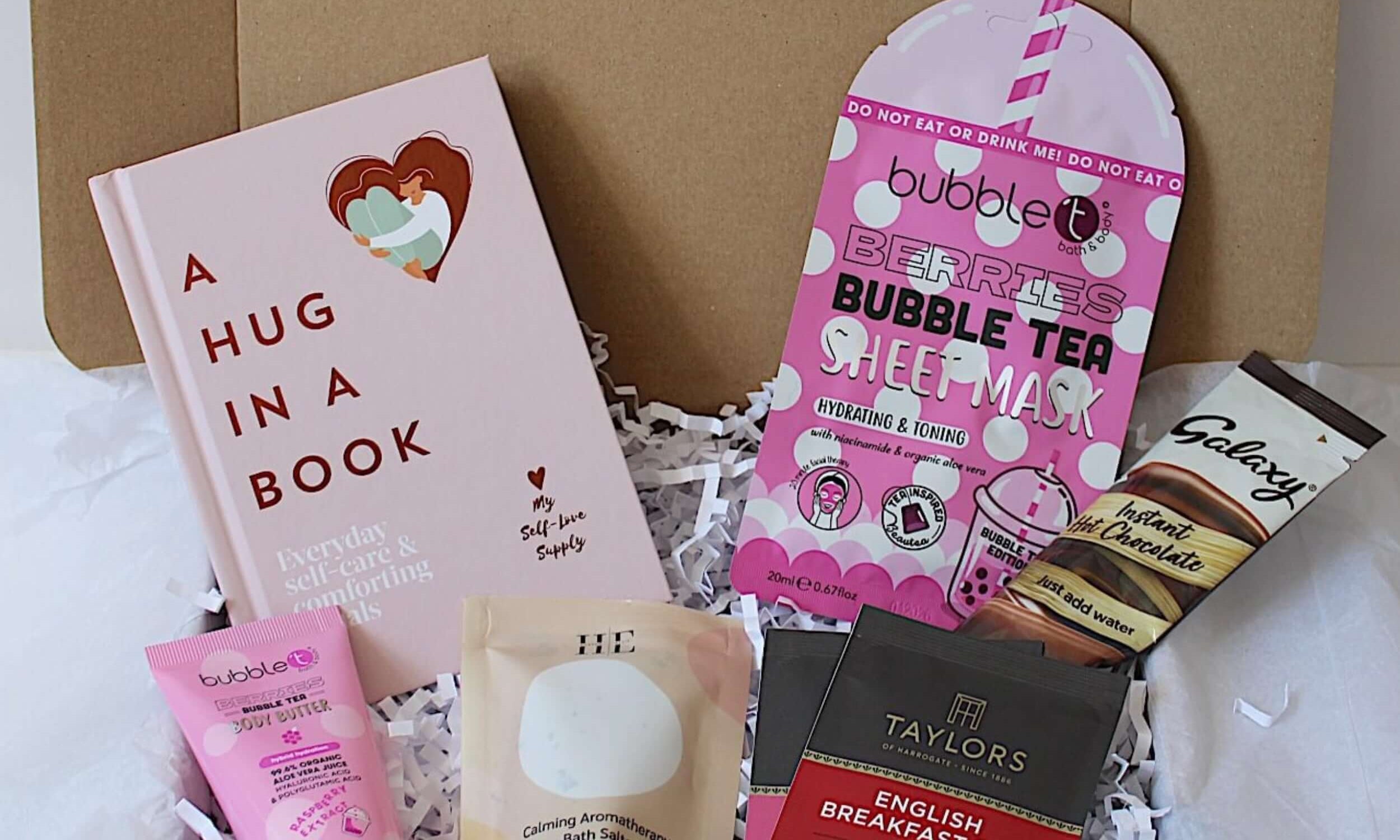 A curated gift box filled with various items including a pink hardcover book titled 'A HUG IN A BOOK', a pink bubble tea sheet mask, berries body butter, aromatherapy bath salts, two individually wrapped tea bags, and a Galaxy chocolate bar, all arranged on white tissue paper inside a gift box.