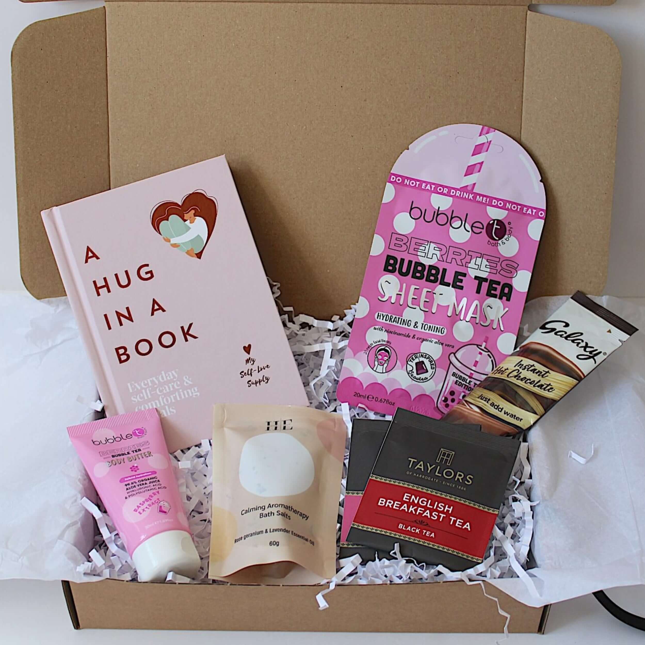 A curated gift box filled with various items including a pink hardcover book titled 'A HUG IN A BOOK', a pink bubble tea sheet mask, berries body butter, aromatherapy bath salts, two individually wrapped tea bags, and a Galaxy chocolate bar, all arranged on white tissue paper inside a gift box.