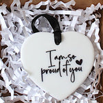 A white heart-shaped ceramic ornament with black text reading 'I am so proud of you' and a small black heart.