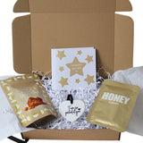 A gift box set containing a white greeting card with gold stars, a gold foil packet of coconut caramel popcorn, a gold foil packet of honeycomb, and a white heart-shaped tag, arranged inside an open gift box with white crinkle paper filler.