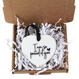 A white heart-shaped ornament with black script reading 'I am so proud of you' sits in a brown cardboard box filled with white shredded paper, with a black ribbon loop at the top.