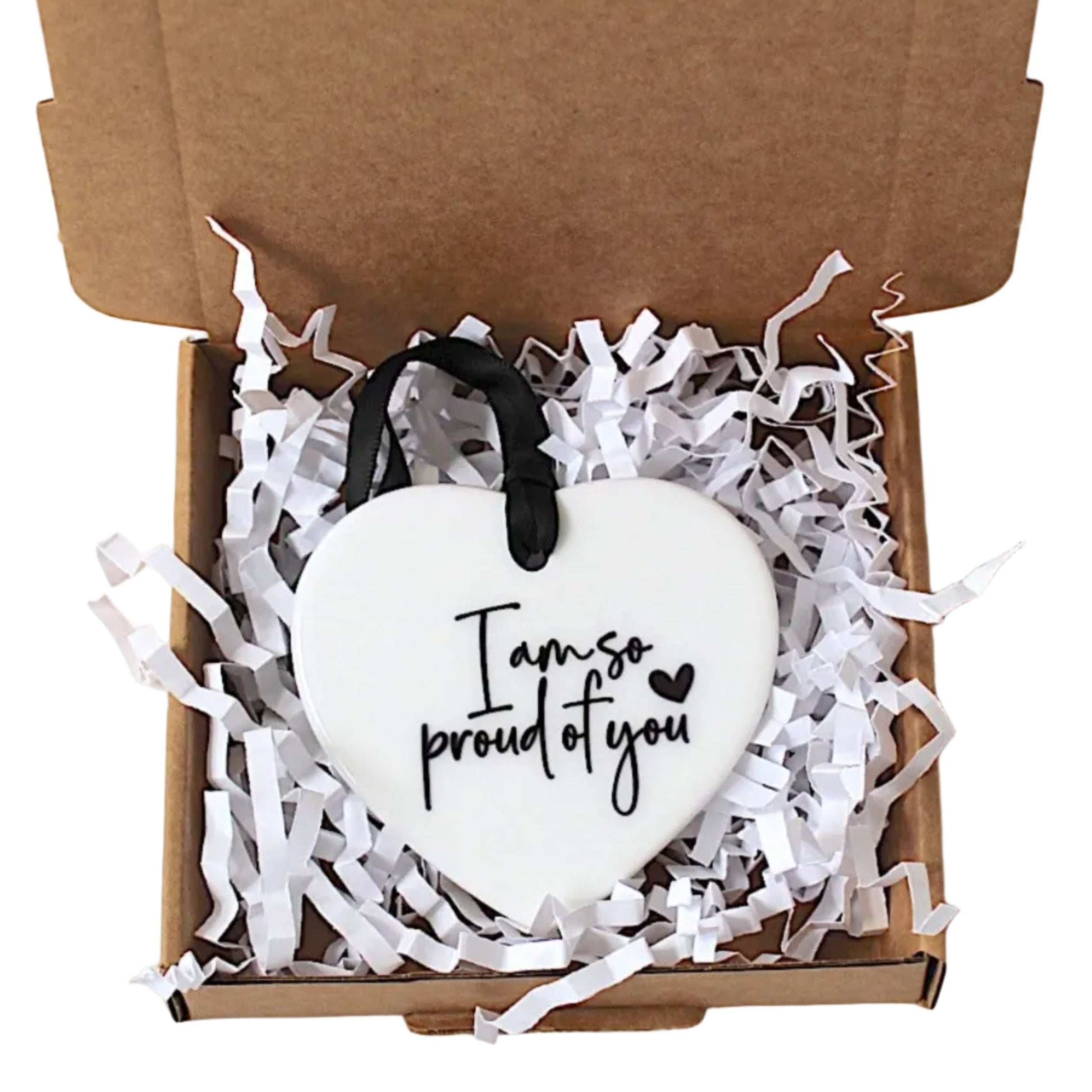 A white heart-shaped ornament with black script reading 'I am so proud of you' sits in a brown cardboard box filled with white shredded paper, with a black ribbon loop at the top.