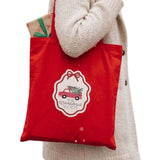 A person wearing a cream knit sweater carries a red fabric tote bag with a white holiday emblem featuring a red car and the text 'its the most wonderful time of the year".