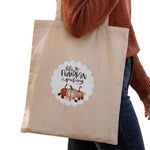 A beige canvas tote bag with a 'Let's go Pumpkin Picking' graphic featuring pumpkins and autumn leaves, carried by a person wearing a rust-colored sweater and dark blue jeans against a white background.