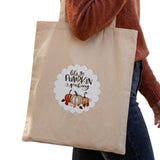 A beige canvas tote bag with a 'Let's go Pumpkin Picking' graphic featuring pumpkins and autumn leaves, carried by a person wearing a rust-colored sweater and dark blue jeans against a white background.