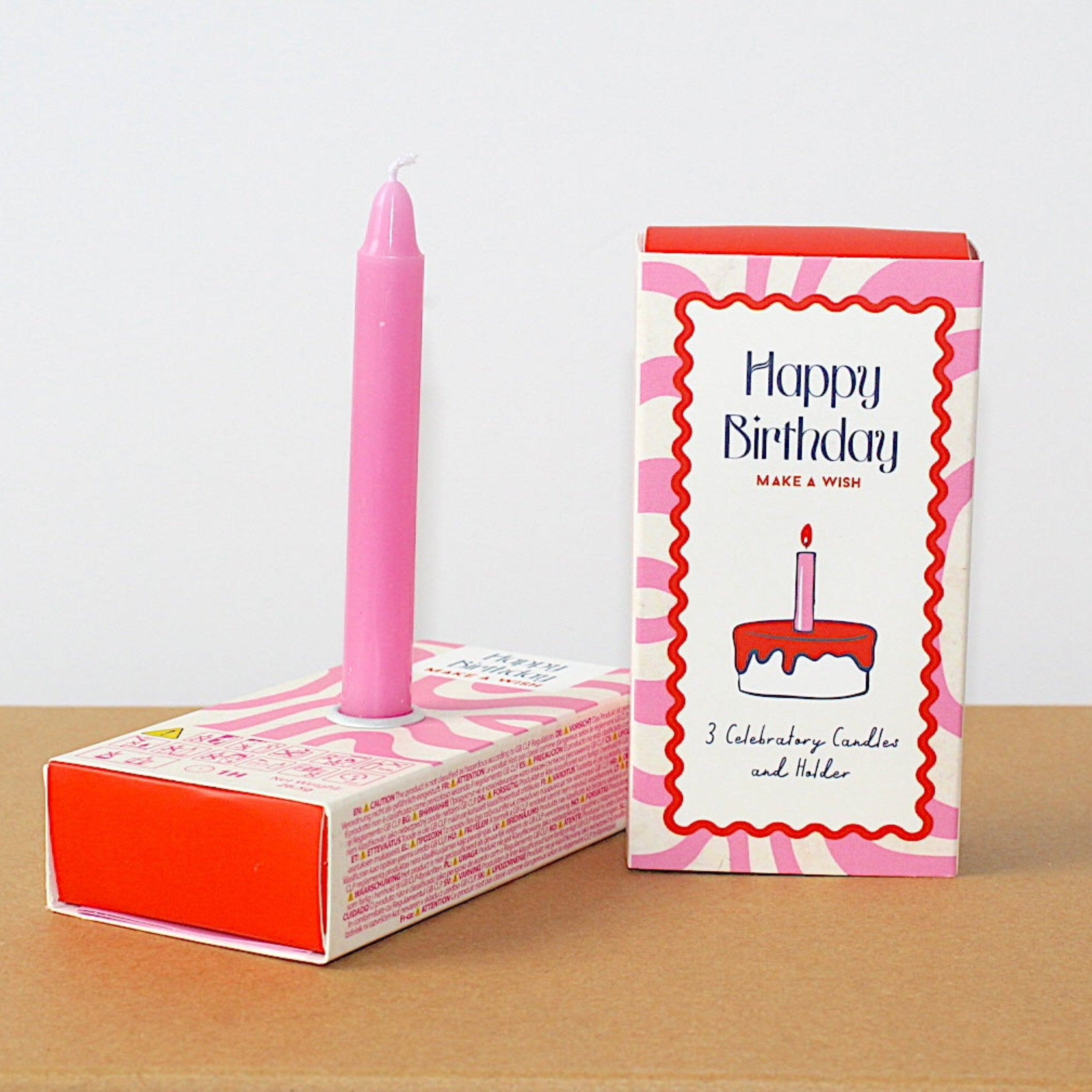 A pink birthday candle standing upright in a pink and white patterned matchbox, with the matching retail box standing beside it on a tan surface against a white background.