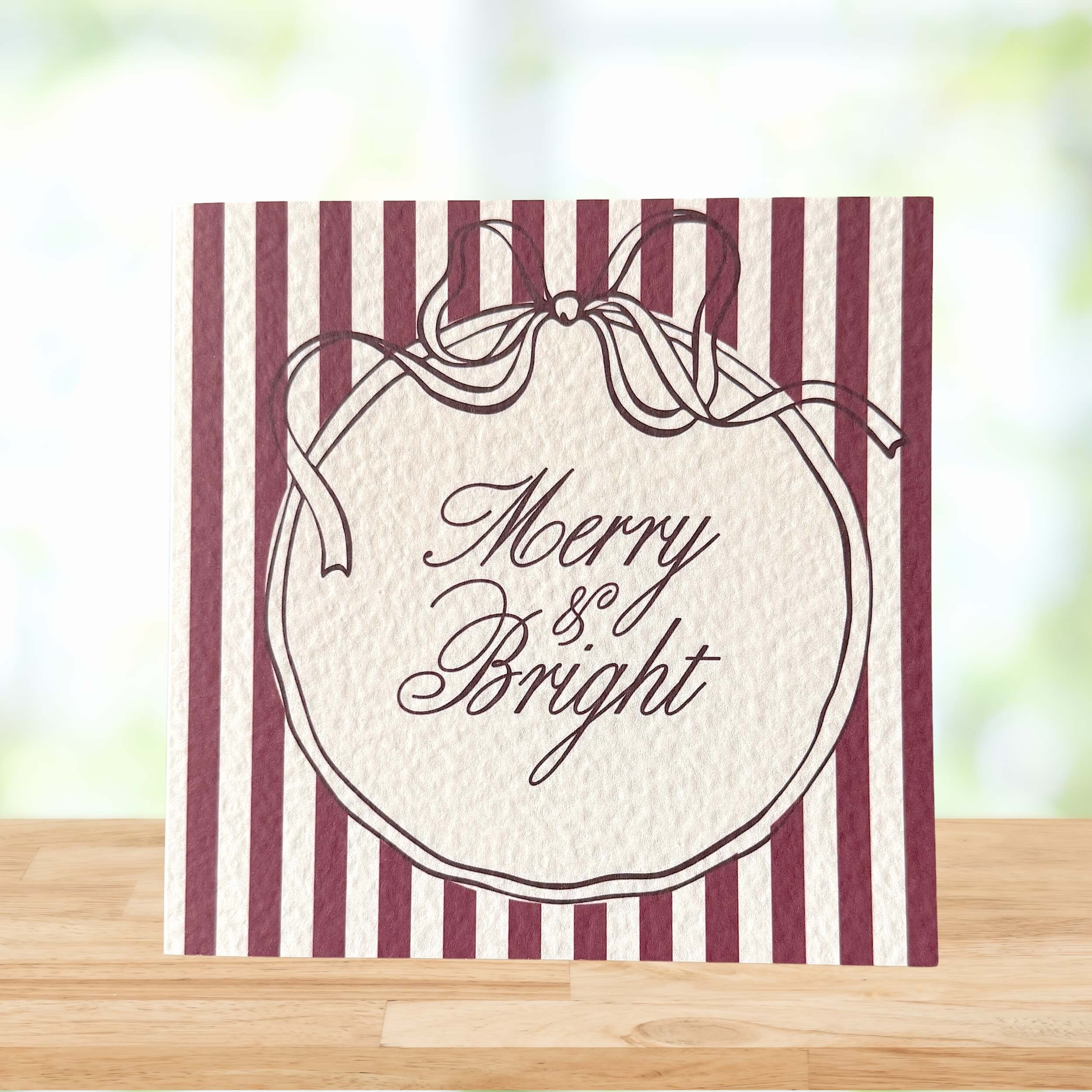 A square greeting card with burgundy and cream vertical stripes, featuring a central cream oval with a ribbon illustration and the words 'Merry & Bright' in script, displayed on a wooden surface with a blurred light background.