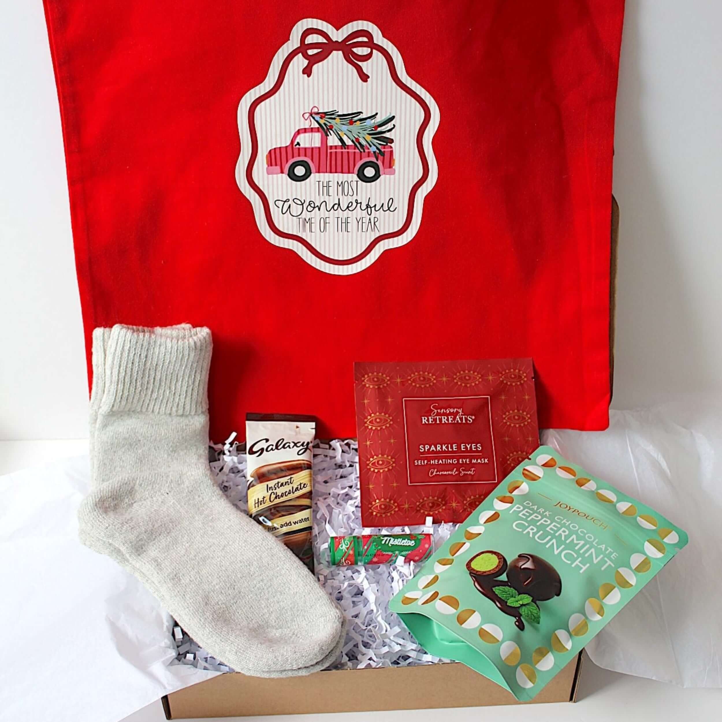 A Christmas-themed gift box set featuring a large red tote bag with a holiday emblem, a light grey knit sock, peppermint crunch sweets,  sensory retreat self heated eye mask, galaxy hot chocolate, candy cane lip balm presented  a gift box with white tissue paper.