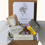 A curated gift box set in a brown magnetic gift box filled with white tissue paper and shredded paper. Visible items include a white card with a bronze line drawing of a woman holding flowers and the text 'Wishing you all, the love this new chapter brings,' 60g aromatherapy bath salts, LSW 'Mind Cards New Munition,'3 individually wrapped 'Taylors Organic tea bags' and a box of 3 soy wax aromatherapy tea lights