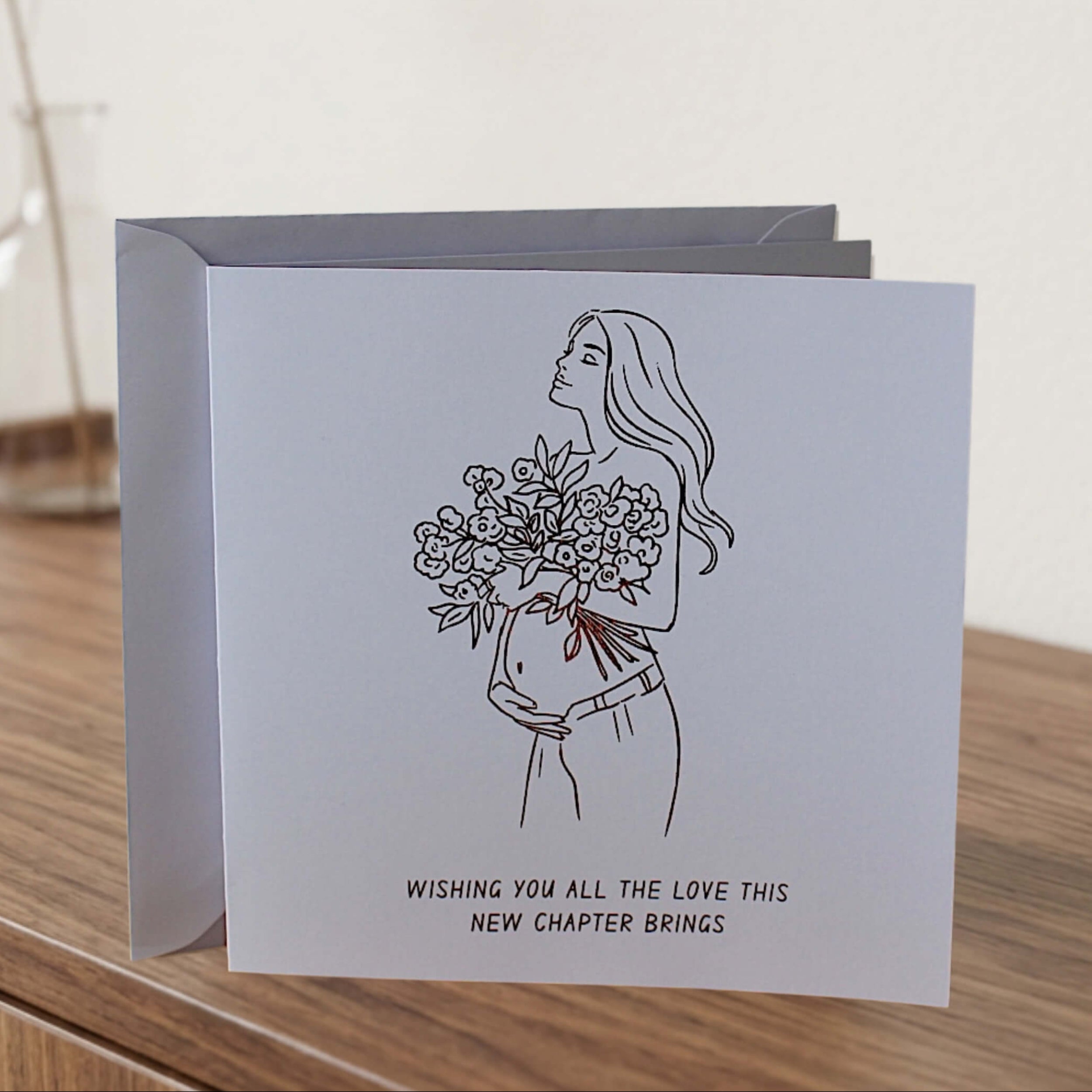 A square greeting card with a bronze foiled  wording and outline of pregnant women holding flowers and a  white envelope, standing on a wooden surface against a light wall. The card features a bronze line drawing of a woman holding a bouquet of flowers, with the text 'WISHING YOU ALL THE LOVE THIS NEW CHAPTER BRINGS' below.