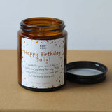 An amber glass soy wax candle jar with a white printed birthday label reading 'Happy Birthday Sally!' and a black lid resting beside it on a kraft paper surface against a light gray background.