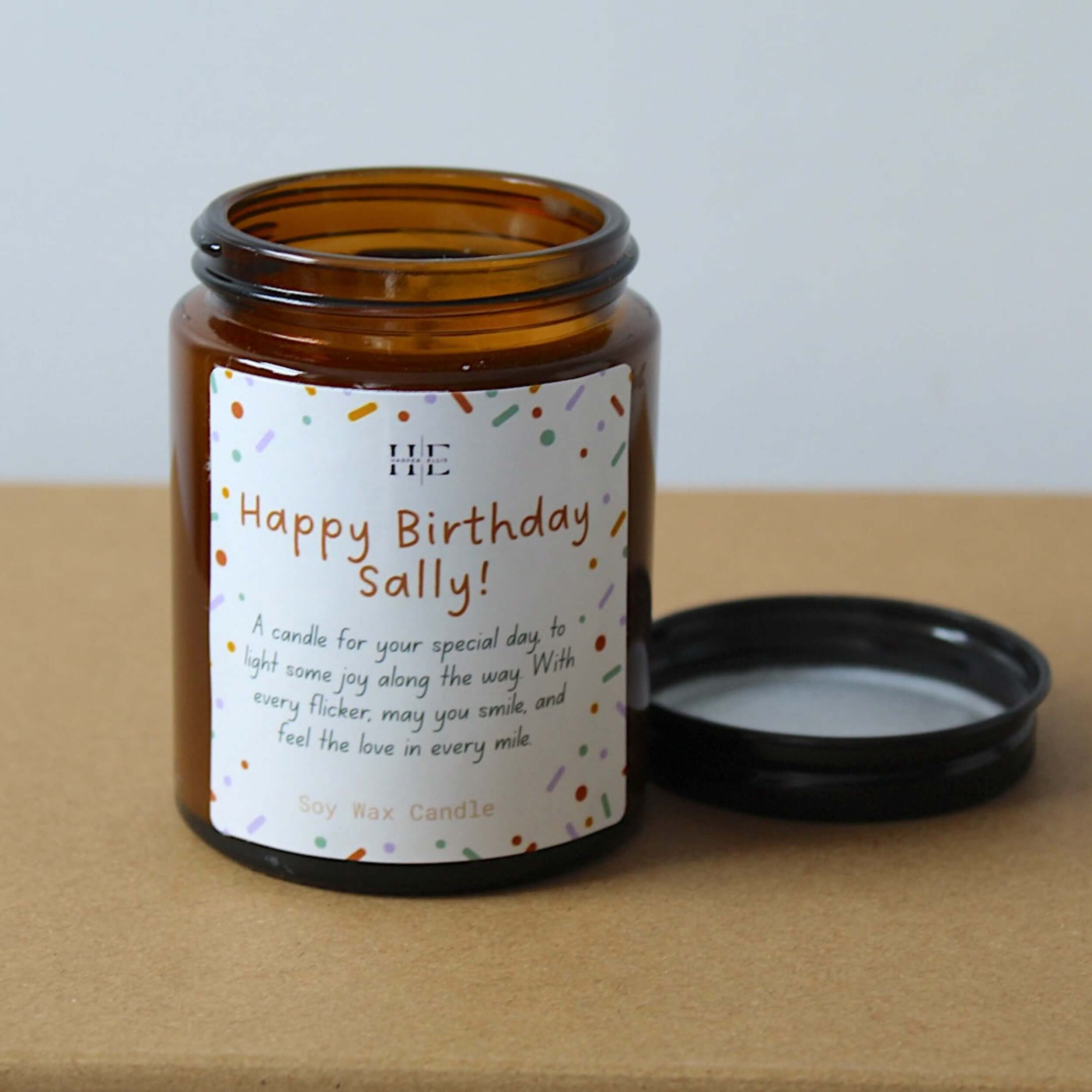 An amber glass soy wax candle jar with a white printed birthday label reading 'Happy Birthday Sally!' and a black lid resting beside it on a kraft paper surface against a light gray background.