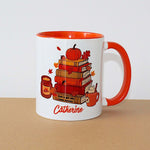A white ceramic mug with a bright orange handle and interior, featuring a fall-themed illustration of stacked books, a pumpkin, leaves, and a mug, with the name 'Catherine' printed in orange script at the bottom. The mug sits on a brown surface against a plain white background.