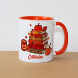 A white ceramic mug with a bright orange handle and interior, featuring a fall-themed illustration of stacked books, a pumpkin, leaves, and a mug, with the name 'Catherine' printed in orange script at the bottom. The mug sits on a brown surface against a plain white background.