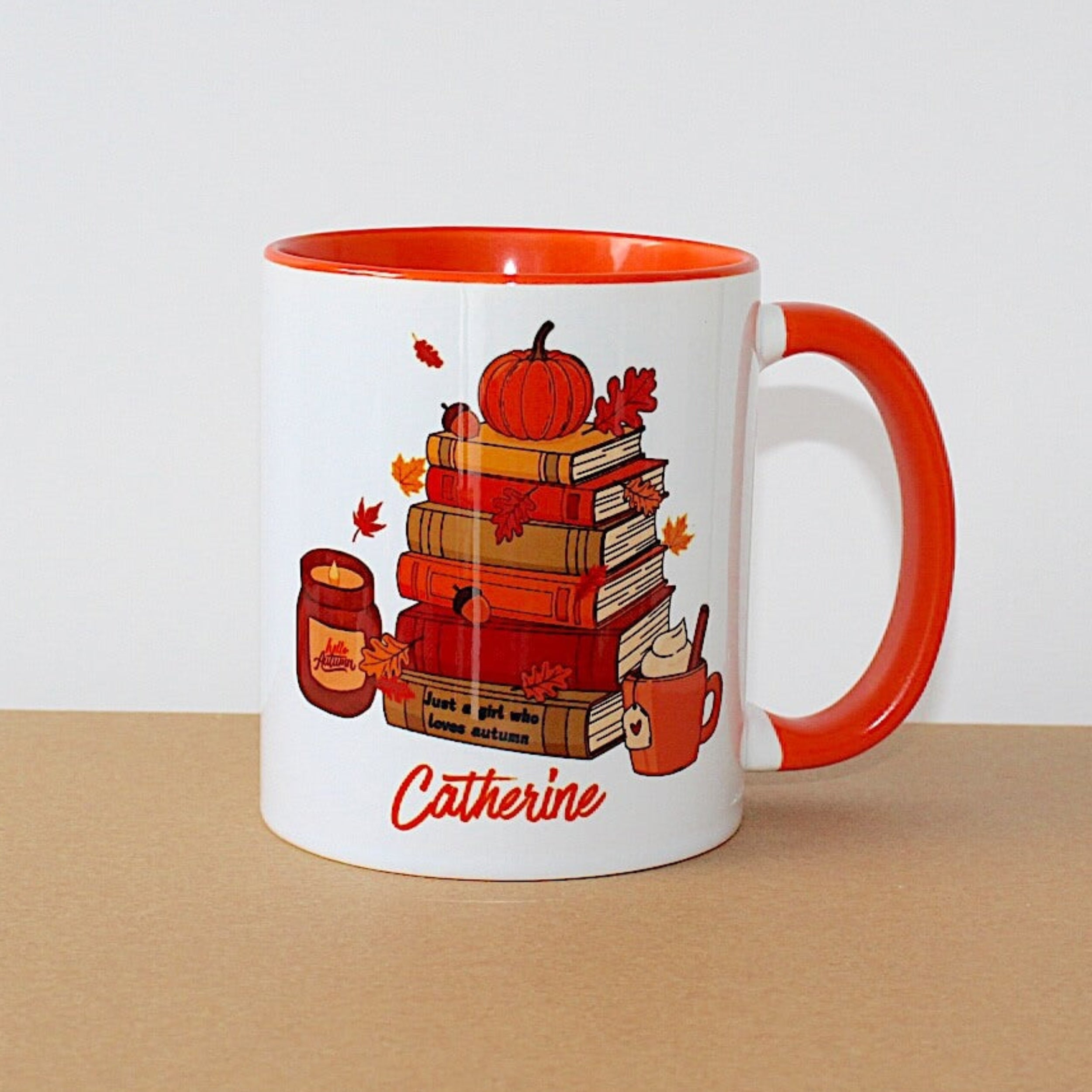 A white ceramic mug with a bright orange handle and interior, featuring a fall-themed illustration of stacked books, a pumpkin, leaves, and a mug, with the name 'Catherine' printed in orange script at the bottom. The mug sits on a brown surface against a plain white background.
