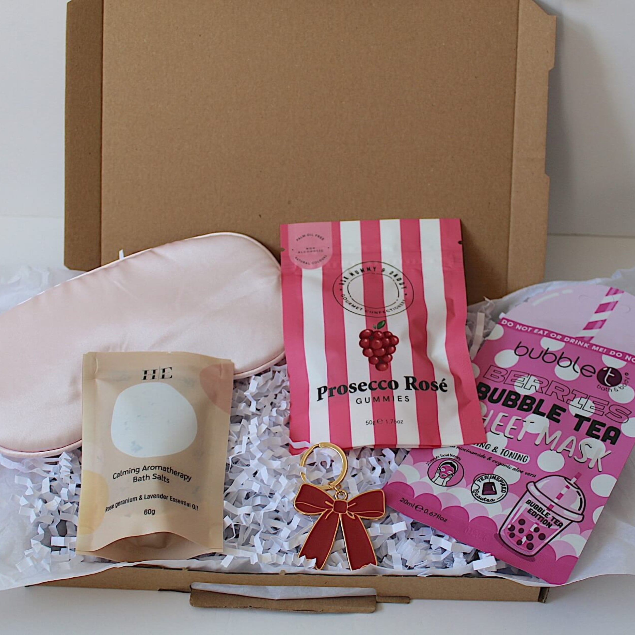 A curated gift box set containing a light pink satin eye mask, aromatherapy bath salts, a pink and white striped Prosecco Rose gummies, red bow metal keyring and a pink Bubble Tea sheet mask arranged in white crinkle paper inside a brown letterbox friendly gift box.