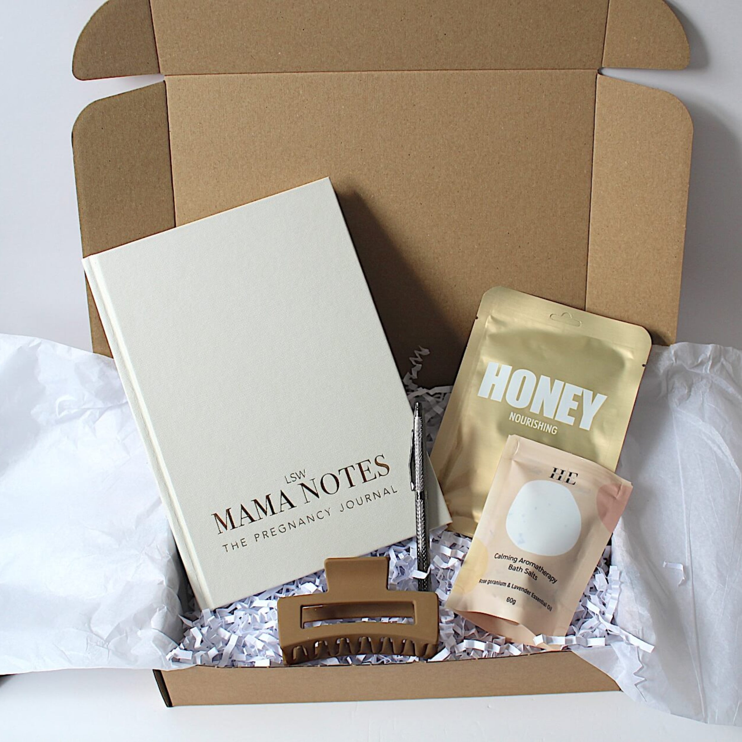 A gift box set containing a cream-colored 'Mama Notes' pregnancy journal, a silver metal pen, a lapcos honey sheet mask, 60g aromatherapy bath salts and a brown hair claw clip, arranged in white tissue paper and crinkle paper inside an open gift box.