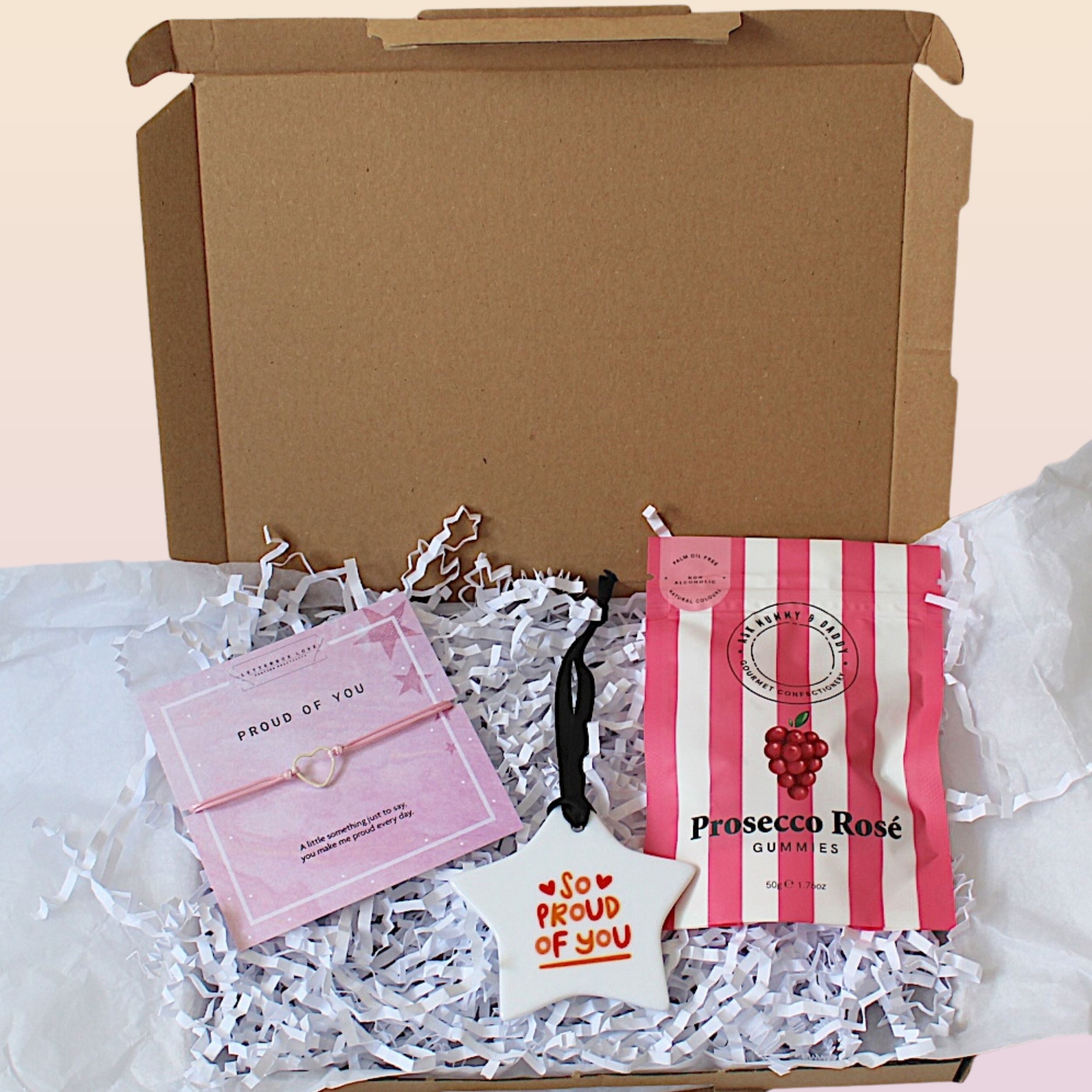 An open gift box filled with white crinkle paper, containing a pink bracelet on a pink card, a white star ornament that says 'So PROUD of you' in red, and a pink-and-white striped bag of Prosecco Rosé Gummies.