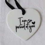 A white heart-shaped ceramic ornament with black script text reading 'I am so proud of you' and a small black heart, hanging from a black ribbon on a white textured fabric background.