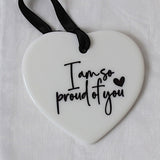 A white heart-shaped ceramic ornament with black script text reading 'I am so proud of you' and a small black heart, hanging from a black ribbon on a white textured fabric background.