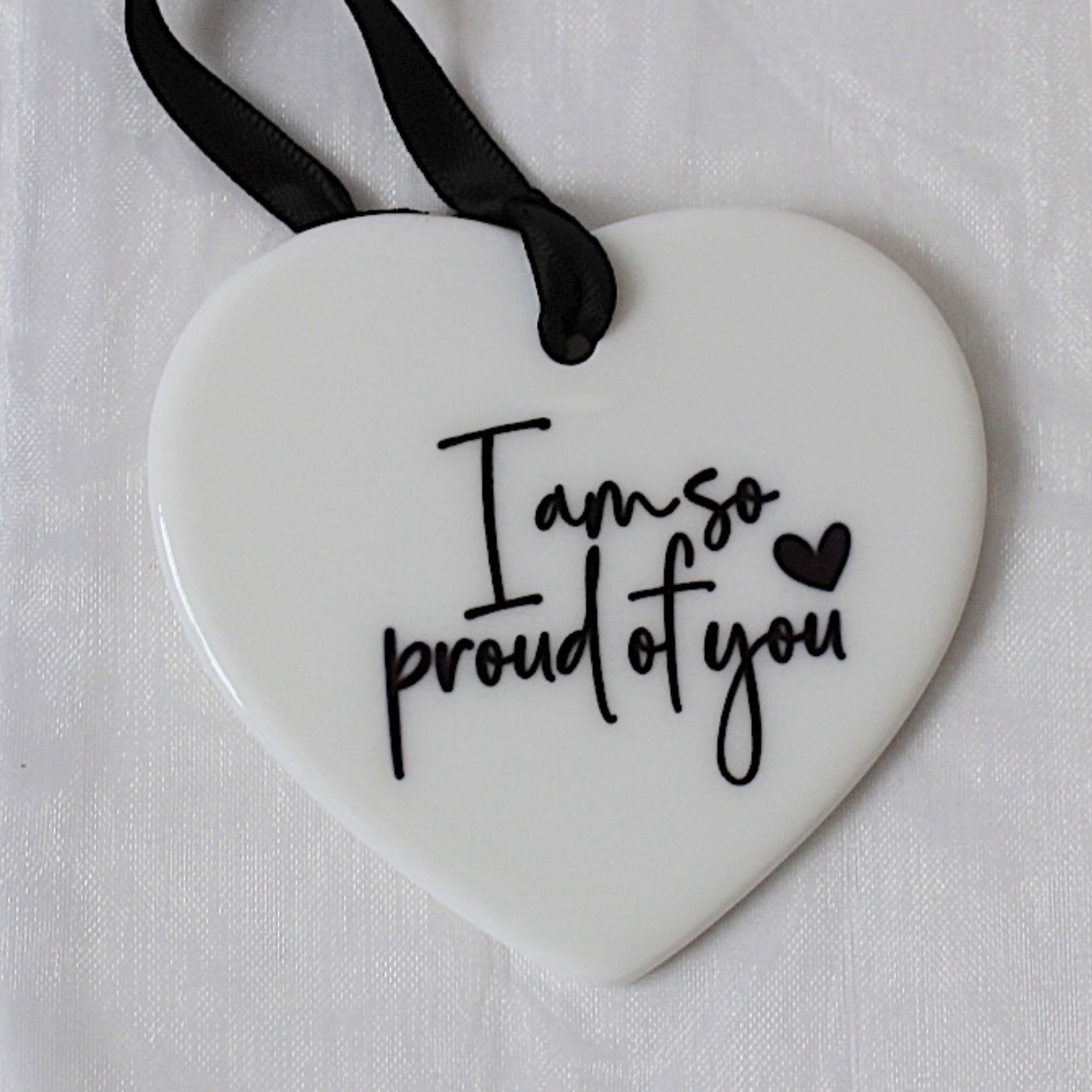 A white heart-shaped ceramic ornament with black script text reading 'I am so proud of you' and a small black heart, hanging from a black ribbon on a white textured fabric background.