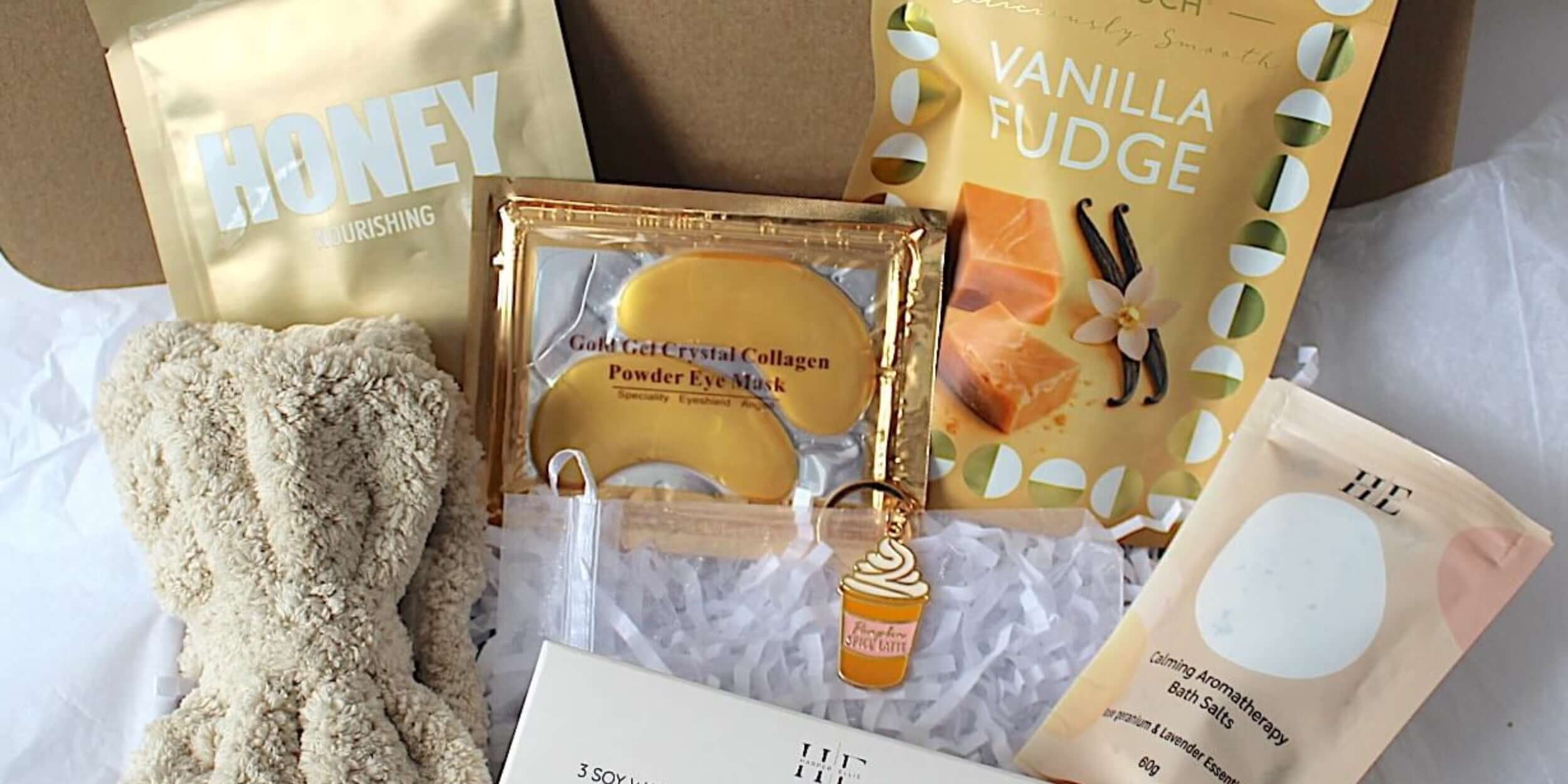 A curated gift box set containing a beige knit beanie, a gold honey pouch, a vanilla fudge snack pouch, a boxed item, a small bottle, and a bath soak packet, arranged in a brown gift box with white tissue paper.