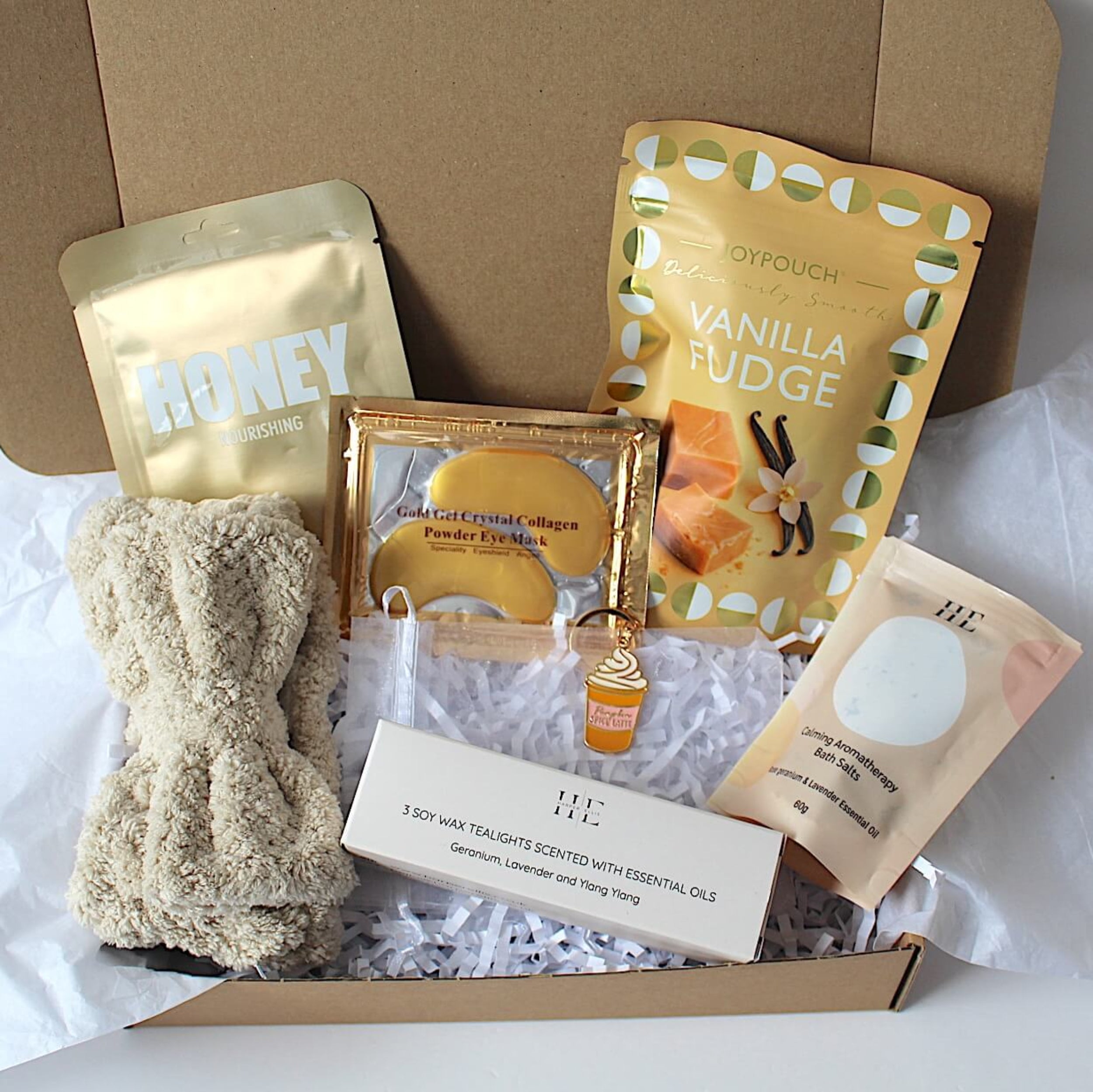 A curated gift box set containing a beige knit beanie, a gold honey pouch, a vanilla fudge snack pouch, a boxed item, a small bottle, and a bath soak packet, arranged in a brown gift box with white tissue paper.