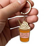 A hand holding a gold-tone metal keychain charm shaped like a cup of spice latte with whipped cream and a straw. The charm has a pink label reading 'Pure Bar Spice Latte' and is shown against a plain white background.