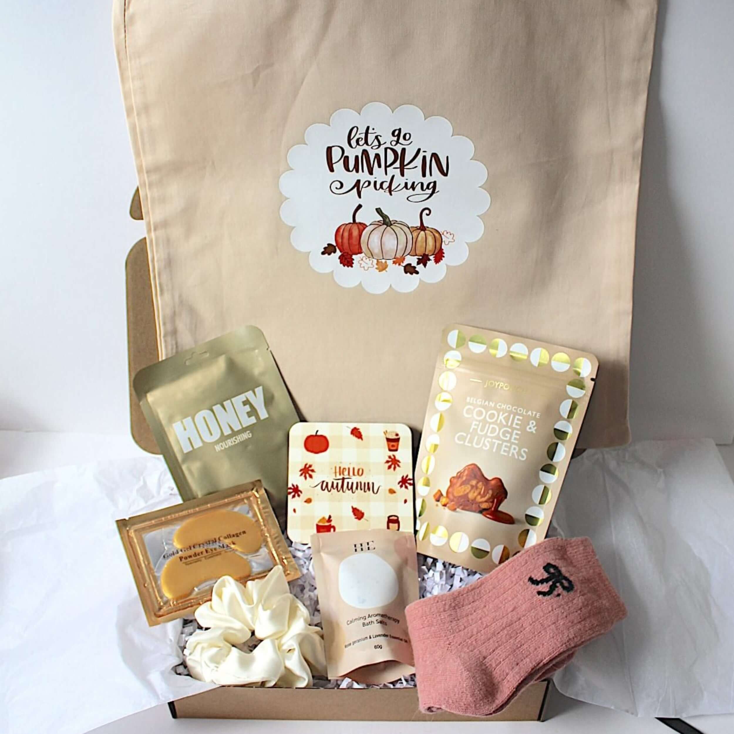 A beige canvas tote bag with a 'Let's Be Pumpkin Munching' graphic, displayed next to a curated gift box filled with fall-themed items including a pink knit sock, cookie and fudge clusters, coaster, under eye patches, aromatherapy bath salts, hair scrunchie  and lapcos honey sheet mask