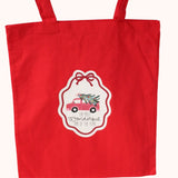A red canvas tote bag with a white and red holiday ornament-shaped graphic in the center, featuring a pink car carrying a Christmas tree and the text 'The most wonderful time of the year'.
