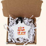 A white star-shaped ornament with the words 'So PROUD of YOU' in orange and red lettering, displayed inside an open brown cardboard gift box filled with white paper shred.