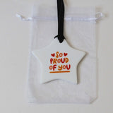 A white star-shaped ornament with the words 'SO PROUD OF YOU' in red and orange lettering, hanging from a black ribbon over a white drawstring pouch on a light background.